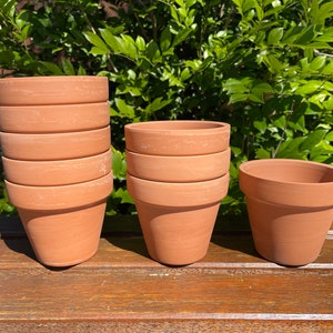 May include: A set of three terracotta flower pots, two stacked and one single, on a wooden surface. The pots are all the same size and shape, with a rounded bottom and a flared lip.
