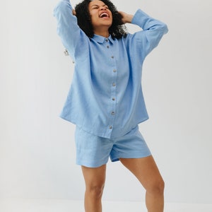 May include: A person wearing a light blue linen shirt and matching shorts. The shirt has a button-up front and a collared neckline. The person is smiling and has their hands on their head.