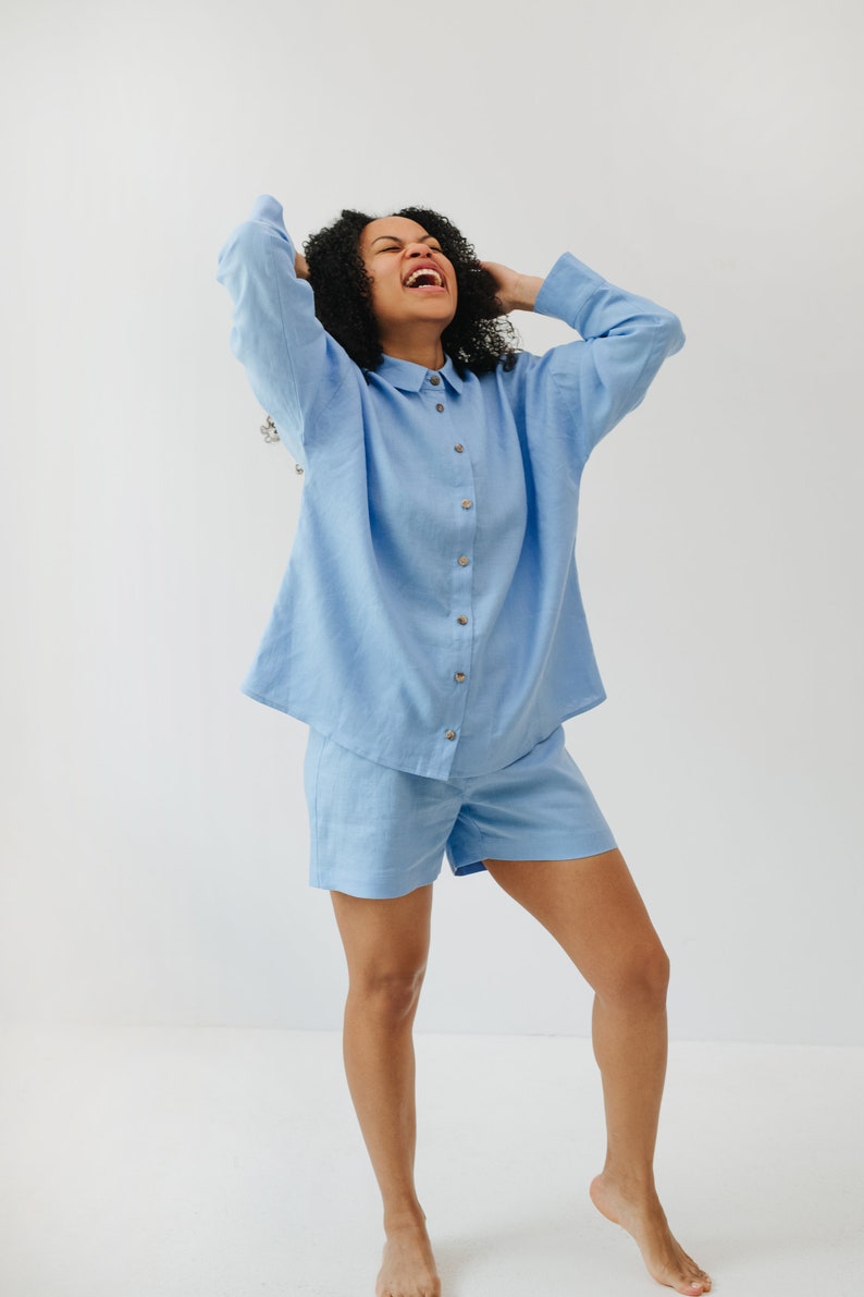 May include: A person wearing a light blue linen shirt and matching shorts. The shirt has a button-up front and a collared neckline. The person is smiling and has their hands on their head.