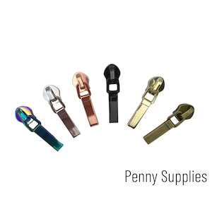 May include: Assortment of six zipper pulls in various colours, including gold, silver, black, rose gold, and iridescent. The zipper pulls are all the same size and shape, with a rectangular base and a small loop at the top.