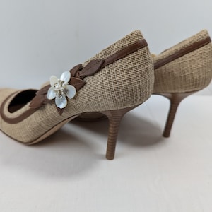 Puede incluir: Un par de zapatos de tac&oacute;n alto tejidos de color beige con ribete de cuero marr&oacute;n y un adorno de flor blanca.