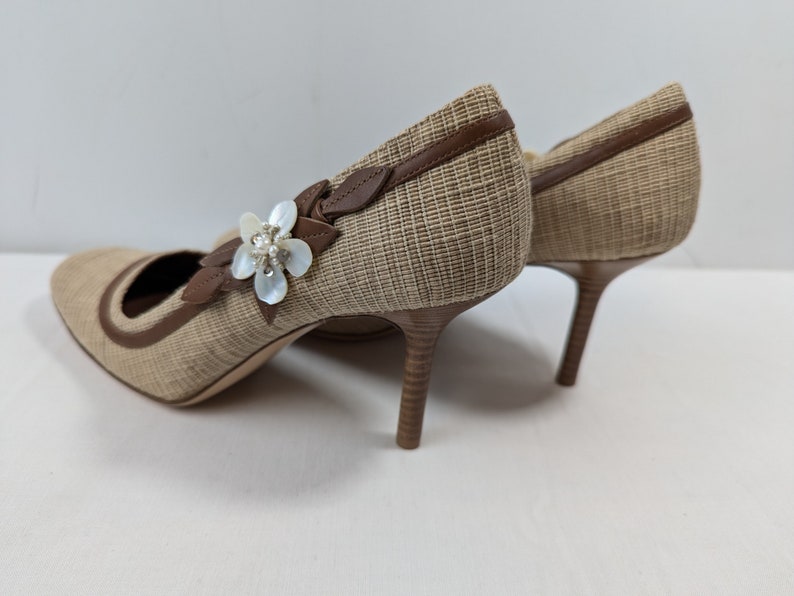 Puede incluir: Un par de zapatos de tac&oacute;n alto tejidos de color beige con ribete de cuero marr&oacute;n y un adorno de flor blanca.