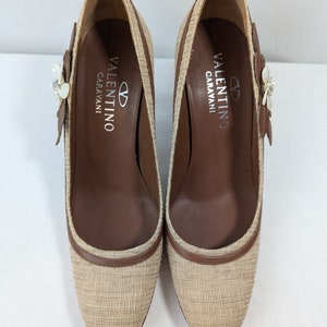 Puede incluir: Un par de zapatos de tac&oacute;n de tejido tejido beige y marr&oacute;n con un adorno floral en la parte superior. Los zapatos tienen el texto "VALENTINO GARAVANI" impreso en el interior.