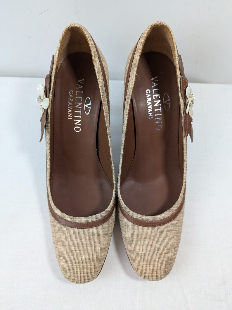 Puede incluir: Un par de zapatos de tac&oacute;n de tejido tejido beige y marr&oacute;n con un adorno floral en la parte superior. Los zapatos tienen el texto "VALENTINO GARAVANI" impreso en el interior.
