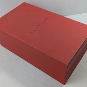 Puede incluir: Una caja de zapatos roja con el texto "VALENTINO GARAVANI" en relieve en la parte superior.