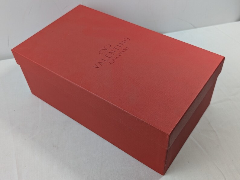Puede incluir: Una caja de zapatos roja con el texto "VALENTINO GARAVANI" en relieve en la parte superior.