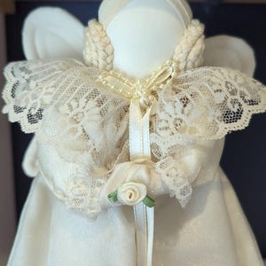 May include: A handcrafted angel doll in ivory and cream hues. The doll has a lace collar, braided hair, and a satin dress. A small white rose with green leaves is on the front. The wings are made of a soft, textured fabric.