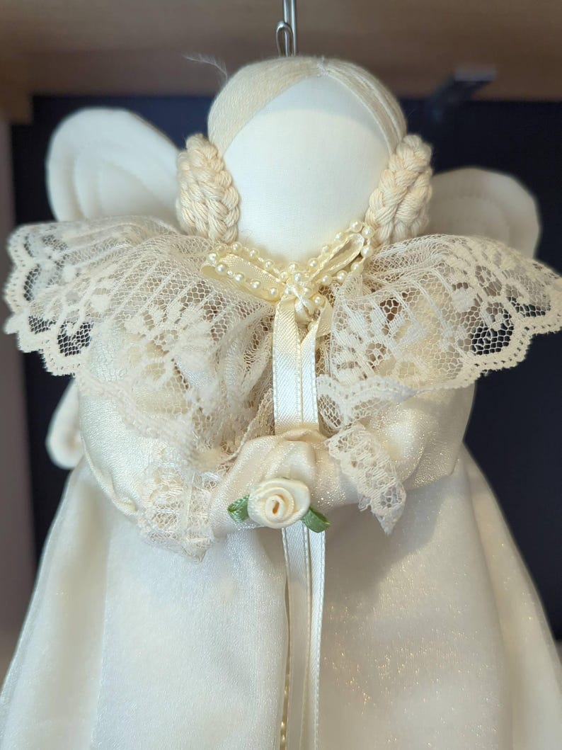 May include: A handcrafted angel doll in ivory and cream hues. The doll has a lace collar, braided hair, and a satin dress. A small white rose with green leaves is on the front. The wings are made of a soft, textured fabric.