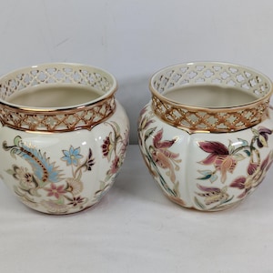 Puede incluir: Dos macetas de cerámica color crema con diseños florales en rosa, azul y dorado. Las macetas tienen un borde dorado decorativo y un borde superior estampado. Las macetas son probablemente para uso decorativo.