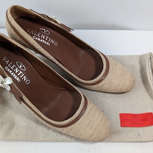Puede incluir: Un par de zapatos Valentino Garavani beige y marr&oacute;n con un adorno floral en la punta. Los zapatos est&aacute;n sobre una superficie de tela beige con una bolsa de polvo Valentino Garavani roja.