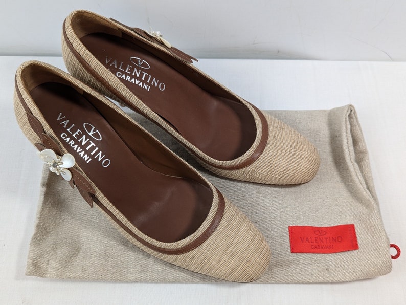 Puede incluir: Un par de zapatos Valentino Garavani beige y marr&oacute;n con un adorno floral en la punta. Los zapatos est&aacute;n sobre una superficie de tela beige con una bolsa de polvo Valentino Garavani roja.