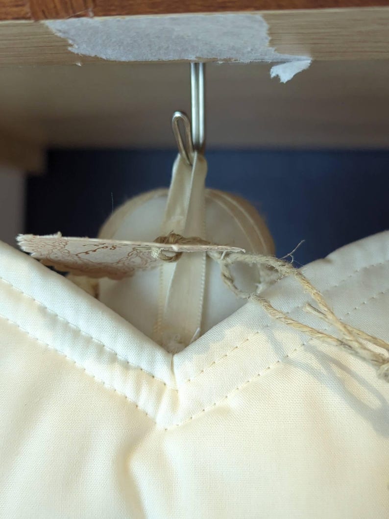 May include: Close-up of a cream-coloured garment with a V-neck, hanging from a metal hook. A tag with a decorative edge is attached with twine. The garment is made of a soft fabric with visible stitching.