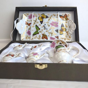 May include: A butterfly-themed tea set in a brown box. The set includes a rectangular tray, a teapot, cups, saucers, and spoons. The tray and tea set are white with butterfly and floral designs, with gold accents.