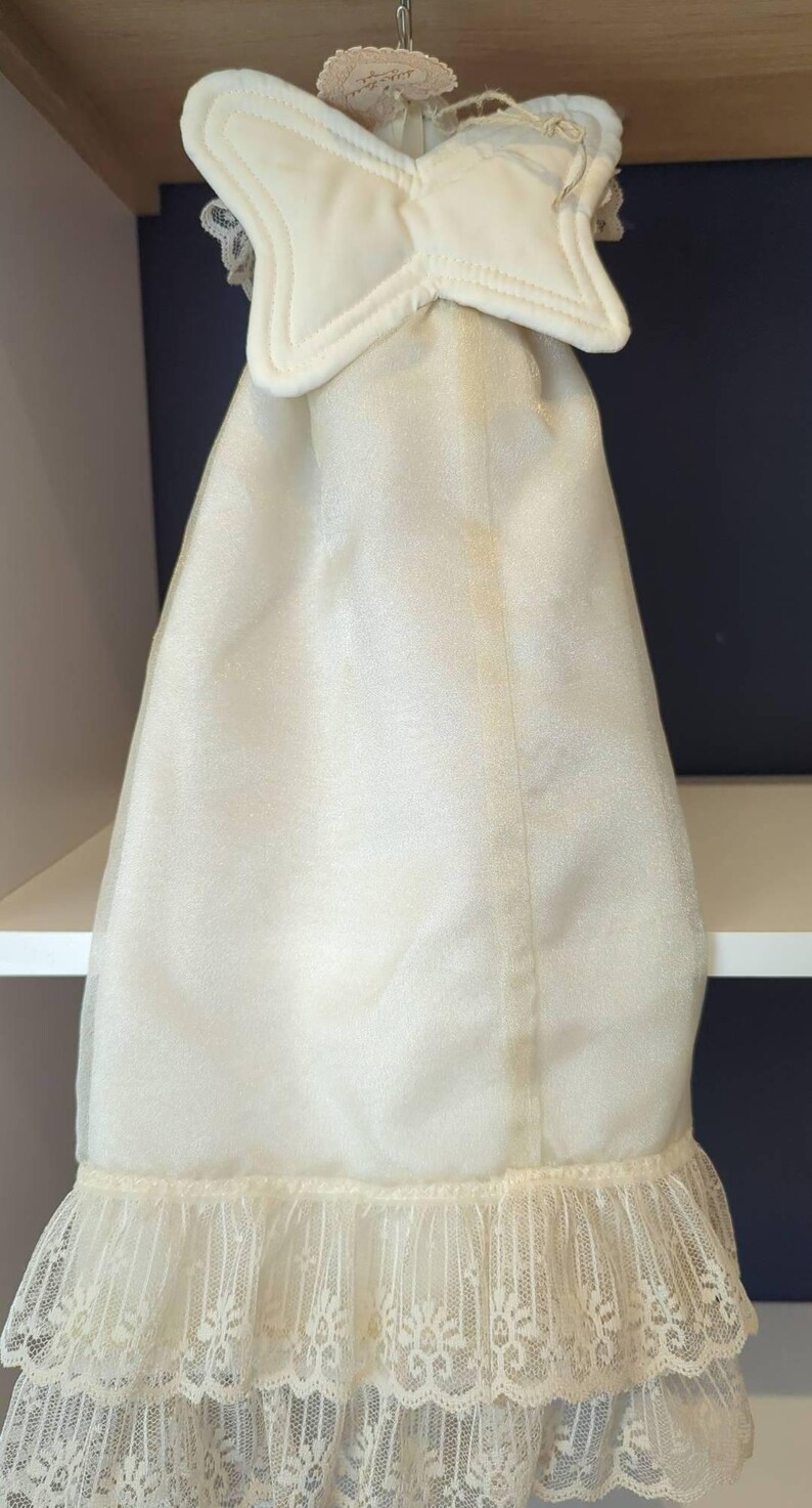 May include: Cream-coloured garment cover with a star-shaped top and a ruffled lace hem. The cover is made of a sheer, flowing fabric. The top has a padded, star-shaped design. The bottom edge features a delicate lace trim.