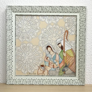 May include: A framed painting depicting the Nativity scene with Mary, Joseph, and baby Jesus. The painting is set against a background of white snowflakes on a beige background. The painting is framed in a white frame with ornate detailing.
