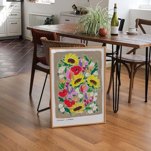 Op de afbeelding: Een ingelijste kunstprint met een kleurrijk bloemstuk met zonnebloemen en klaprozen. Het kunstwerk heeft een neutrale achtergrond en staat op een houten vloer. De tekst "flower market" is onderaan te zien.