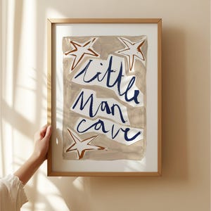 Op de afbeelding: Een ingelijst afdruk met een beige achtergrond en blauwe tekst die "little man cave" luidt. De afdruk bevat drie bruine sterren.
