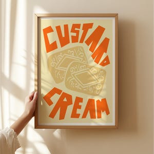 May include: A framed print with a yellow background and orange text that reads "Custard Cream". The print features a stylized illustration of two custard cream biscuits.