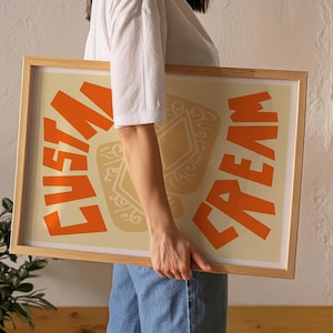 May include: A person holding a framed print with the words "Custard Cream" in orange letters on a cream background. The print features a stylized illustration of a custard cream biscuit.