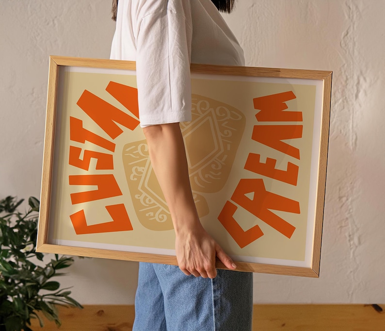 May include: A person holding a framed print with the words "Custard Cream" in orange letters on a cream background. The print features a stylized illustration of a custard cream biscuit.