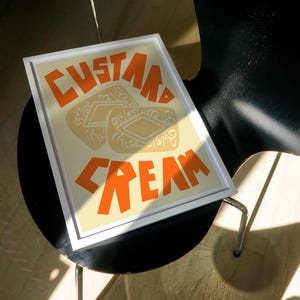 May include: A framed print with a white background and orange text that reads "Custard Cream". The image features three custard cream biscuits.