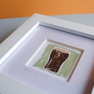 May include: A framed artwork featuring a brown beer glass illustration on a green background. The artwork is in a white frame and includes the text "Split the G. N.C."