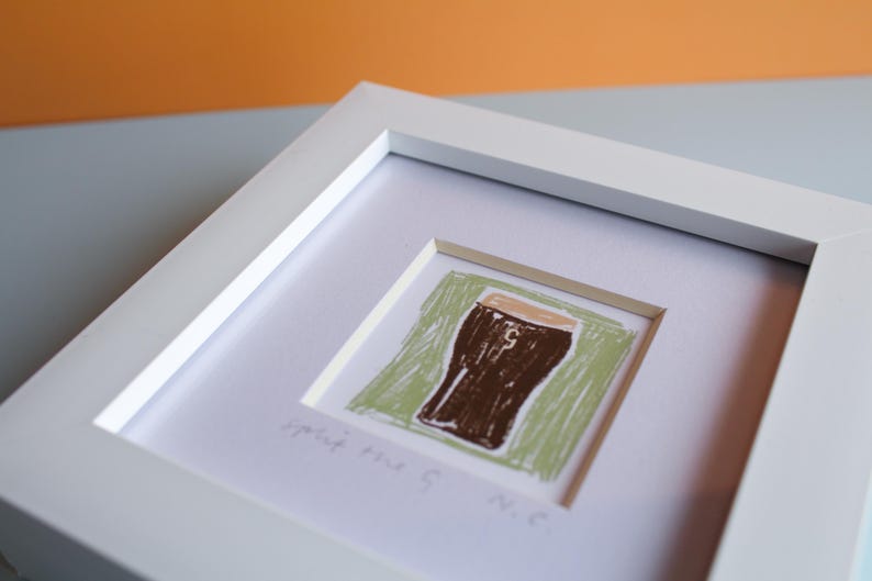 May include: A framed artwork featuring a brown beer glass illustration on a green background. The artwork is in a white frame and includes the text "Split the G. N.C."