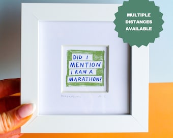May include: A square white frame containing a small print with a green textured background and blue handwritten text that reads: DID I MENTION I RAN A MARATHON? The print is centred within a white mat board and includes a small signature.