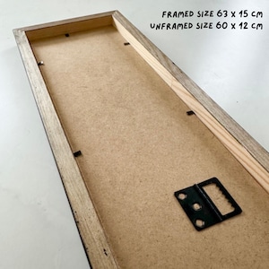 May include: The back of a framed print with a wooden frame. The frame has a black metal hanging hook. The text on the frame reads "FRAMED SIZE 63 x 15 cm UNFRAMED SIZE 60 x 12 cm".