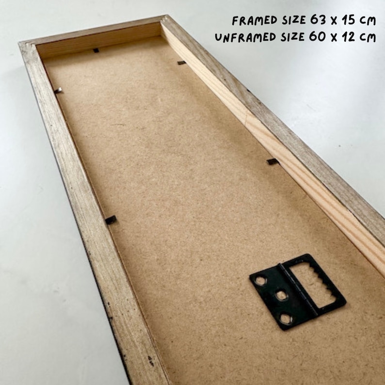 May include: The back of a framed print with a wooden frame. The frame has a black metal hanging hook. The text on the frame reads "FRAMED SIZE 63 x 15 cm UNFRAMED SIZE 60 x 12 cm".