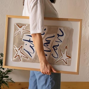 Op de afbeelding: Een ingelijst drukwerk met een beige achtergrond en blauwe en bruine abstracte vormen. De print bevat de tekst "life is now" en verschillende sterren.