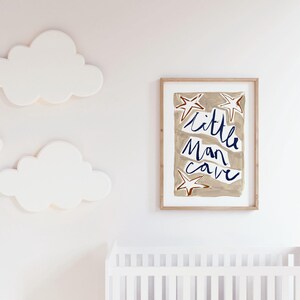 Op de afbeelding: Een ingelijst print met een beige achtergrond en witte sterren. De tekst "little man cave" is geschreven in blauwe cursieve letters.