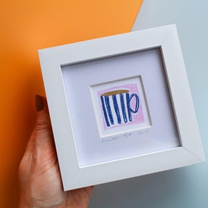May include: A framed art print depicting a mug with blue vertical stripes and a gold rim on a pink background. The artwork is in a white square frame. The background is a mix of orange and pale blue.