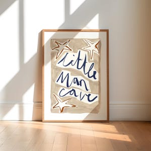 Op de afbeelding: Een ingelijst print met een beige achtergrond en blauwe tekst die "little man cave" luidt. De print heeft vier bruine sterren.
