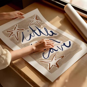Op de afbeelding: Een wit canvas met een beige achtergrond en blauwe letters die "little cave" lezen. Het canvas is versierd met bruine stervormen.
