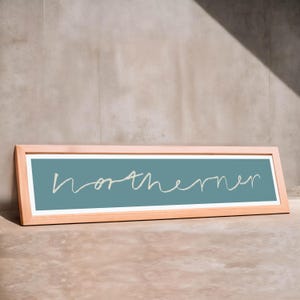 May include: A rectangular wooden framed sign with the word "northerner" in white cursive script on a teal background. The frame is a light wood color, and the sign is leaning against a neutral-colored wall.