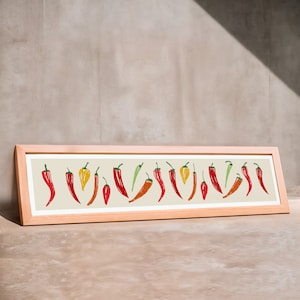 May include: A framed art print featuring a row of stylized chili peppers in various colors, including red, orange, yellow, and green, against a beige background. The frame is a light wood color, and the artwork is displayed on a neutral surface.
