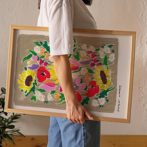 Op de afbeelding: Een ingelijste bloemenkunstprint met een boeket zonnebloemen, klaprozen en andere bloemen in geel, rood, roze en wit. Het kunstwerk is geplaatst tegen een neutrale achtergrond en ingelijst in licht hout.