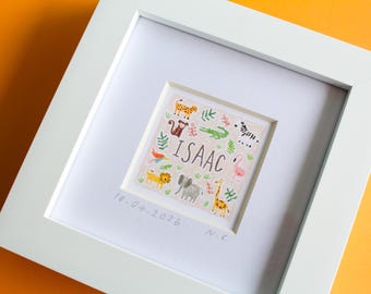 May include: A white framed artwork with a square illustration. The illustration features the name "ISAAC" and drawings of animals. The date "10-04-2026" is written below the artwork. The frame is white, set against a white background.