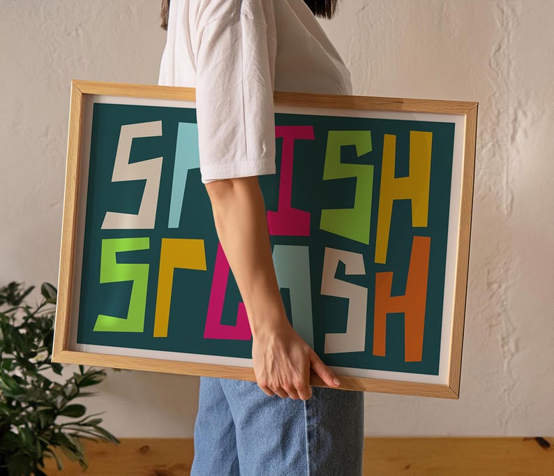May include: A framed art print with the word "SPLASH" in colorful, bold, block letters. The letters are in white, light blue, pink, green, orange, and yellow against a teal background. The frame is made of light-colored wood.