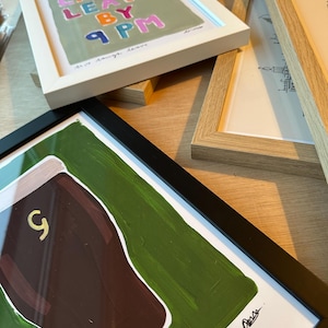 May include: A selection of framed art prints. One print, in a white frame, displays the text "Live Laugh Leave By 9 PM" in various colours. Another, in a black frame, shows a brown shape with a gold '9' on a green background.