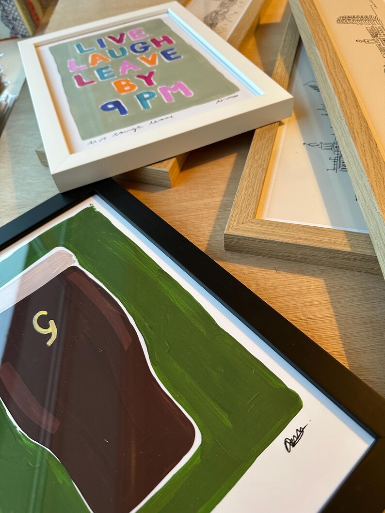 May include: A selection of framed art prints. One print, in a white frame, displays the text "Live Laugh Leave By 9 PM" in various colours. Another, in a black frame, shows a brown shape with a gold '9' on a green background.