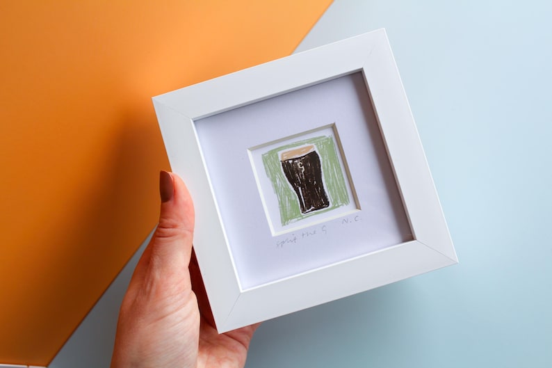 May include: A white framed artwork with a drawing of a pint of stout. The artwork has a white mount. The drawing is on a green background and has the text "spring time 9 N.C." at the bottom.
