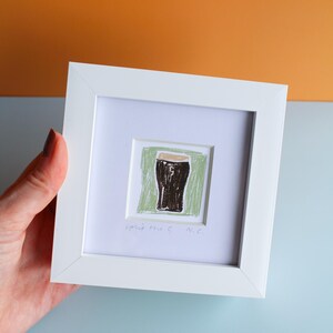 May include: White framed artwork featuring a drawing of a dark stout in a glass, set against a green background. The artwork includes the text "spirit the G N.C." The frame is held against a backdrop of orange and blue.