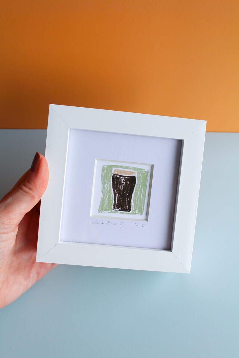 May include: White framed artwork featuring a drawing of a dark stout in a glass, set against a green background. The artwork includes the text "spirit the G N.C." The frame is held against a backdrop of orange and blue.
