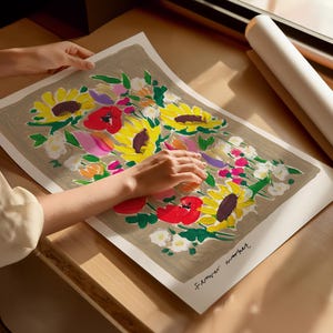 Op de afbeelding: Een bloemenkunstprint met zonnebloemen, klaprozen en andere bloemen in geel, rood, roze en paars. Het kunstwerk staat op een neutrale achtergrond en is gesigneerd. Op de achtergrond is een opgerolde print te zien.