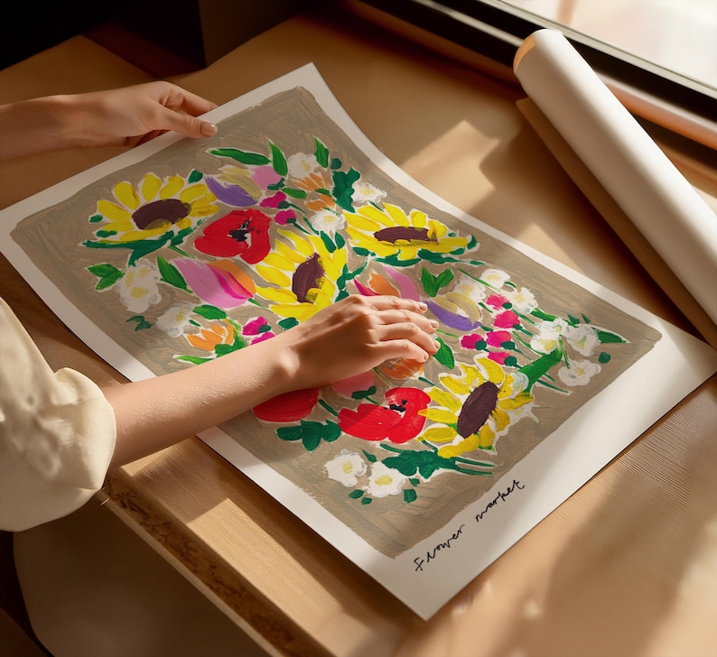 Op de afbeelding: Een bloemenkunstprint met zonnebloemen, klaprozen en andere bloemen in geel, rood, roze en paars. Het kunstwerk staat op een neutrale achtergrond en is gesigneerd. Op de achtergrond is een opgerolde print te zien.