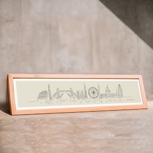 May include: A framed black and white line drawing of the London skyline, including the London Eye, Big Ben, and the Shard. The drawing is on a beige background and the frame is light wood.