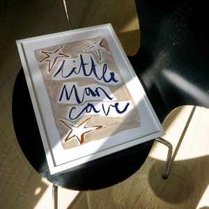 Op de afbeelding: Een wit ingelijst print met een beige achtergrond en blauwe letters die "little man cave" zeggen. Er zijn drie bruine sterren op de print.