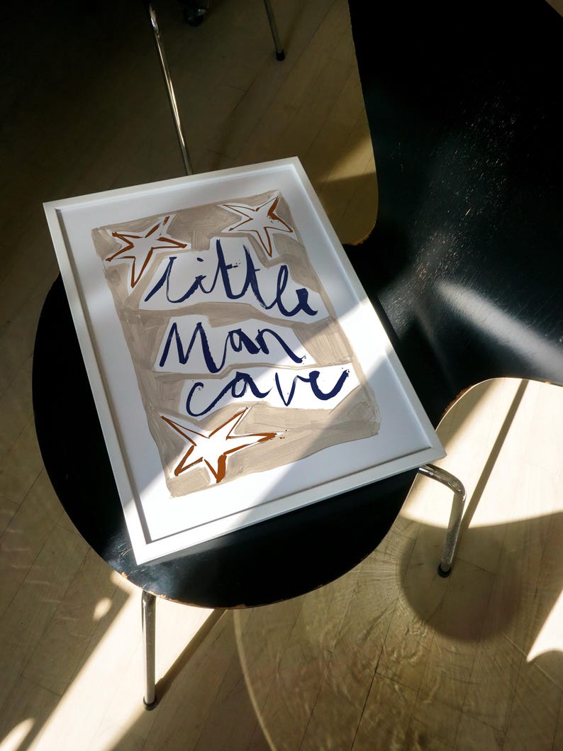 Op de afbeelding: Een wit ingelijst print met een beige achtergrond en blauwe letters die "little man cave" zeggen. Er zijn drie bruine sterren op de print.